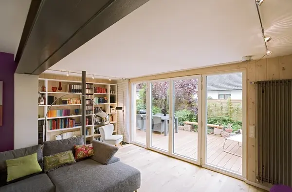 Modernes Wohnzimmer mit Bücherregal, großer Glasfront und Blick auf eine Holzterrasse.
