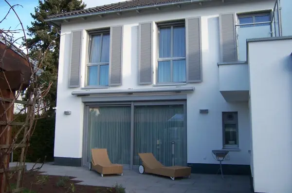 Modernes Einfamilienhaus mit großen Fenstern, Terrasse und Gartenmöbeln im Sonnenschein.
