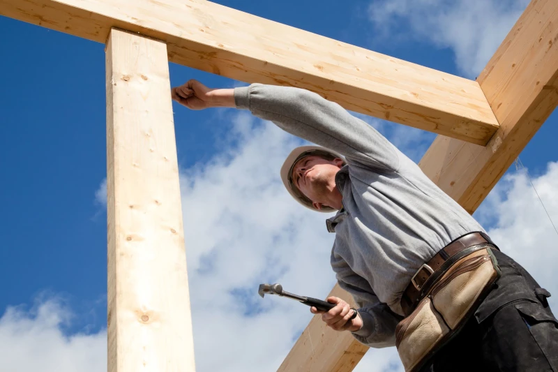Ein Zimmermann baut ein Holzhaus und arbeitet mit Hammer unter blauem Himmel.