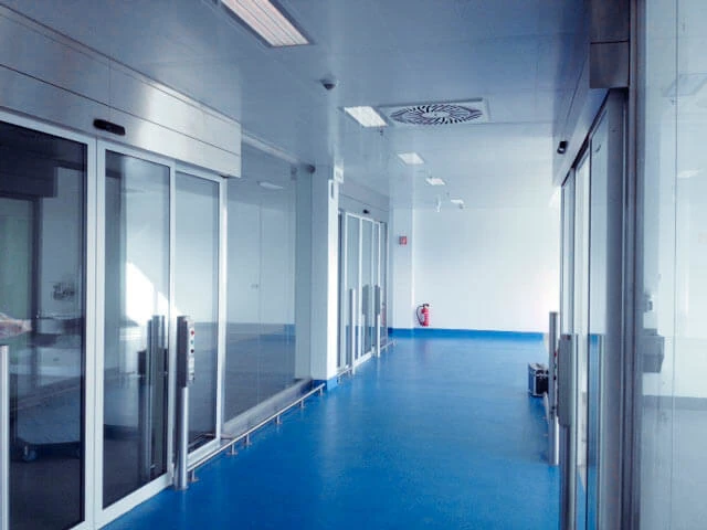 Leerer Klinikflur mit Glas-Schiebetüren, blauem Boden und Feuerlöscher.